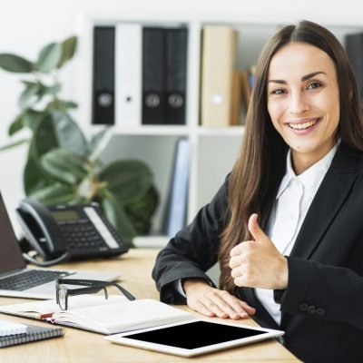 smiling-young-businesswoman-sitting-workplace-showing-thumb-up-sign
